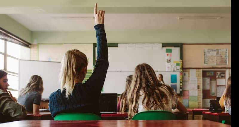 Students in class listening to lecture and raising hands.